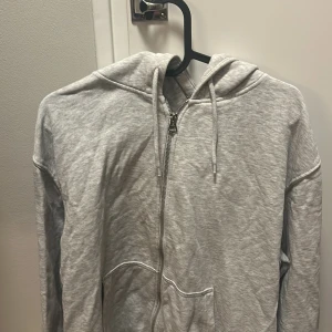 Grå hoodie med dragkedja och huva - Säljer en ljusgrå hoodie med dragkedja framtill och huva med snörning. Tröjan har långa ärmar och en stor ficka på magen. Perfekt för chill dagar och enkel att matcha med jeans eller mjukisbyxor.