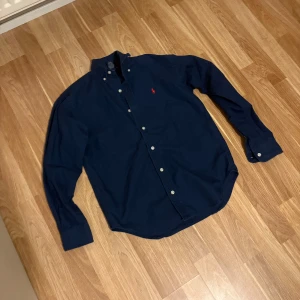 Mörkblå skjorta från Ralph Lauren - Klassisk mörkblå skjorta från Ralph Lauren i 100% bomull. Skjortan har button-down krage, vita knappar och den ikoniska röda loggan broderad på bröstet. Långärmad modell med normal passform, perfekt för en clean och stilren look.
