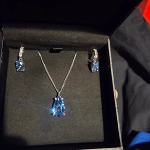 Set med blått halsband och örhängen - Snyggt smyckeset med ett halsband och matchande örhängen i silverkedja. Med blå ädelstenar som huvudsten på både örhängen och halsband med kristall på örhängena. Den reflekterar ljusblå väldigt bra och glänser snyggt. Änvänd 1 gång endast. Köpt på halbergs guld. Rengöringsduk för stenarna medföljer.