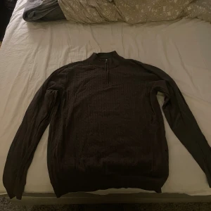 Mörkbrun stickad half zip tröja - Snygg mörkbrun stickad tröja med half zip och hög krage. Tröjan har ett diskret mönster och långa ärmar, perfekt för kyliga dagar. Passar dig som gillar klassisk stil med en modern twist.