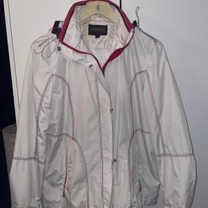 Vit vindjacka från Mode Mille Design - Snygg vit vindjacka med röda och blå detaljer från Mode Mille Design. Jackan har huva, dragkedja framtill och coola kontrastsömmar. Perfekt för dig som gillar sportig stil och vill ha något som sticker ut lite extra. Passar dig som vill ha en jacka med lite edge.