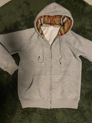 Grå hoodie från Burberry med rutig huva - Snygg grå hoodie från Burberry i storlek   L  passar  storlek S med klassisk rutig huva i beige, svart och röd. Tröjan har dragkedja, snörning och två fickor framtill. Materialet är mjuk bomull och huvan har metalländar med Burberry-logga. Perfekt för dig som gillar stilrena detaljer.