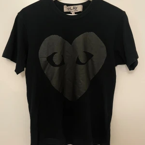 Svart Comme des Garçons t-shirt - Svart t-shirt från Comme des Garçons (CDG) med stort hjärtmotiv med ögon på framsidan och ett hjärta på baksidan. Materialet är mjuk bomull som känns skön mot huden. Väldigt fint skick och sparsamt använd, är i storlek S men sitter lite mindre xs/s skulle jag mer säga! Hör av dig vid minsta lilla 🤗🤩
