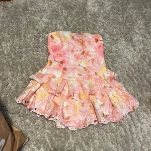 Blommig rosa kortklänning med volanger - Superfin axelbandslös kortklänning med blommigt mönster i rosa, gult och vitt. Klänningen har flera lager volanger med broderade håldetaljer och en söt, romantisk vibe. Perfekt för dig som gillar färg och detaljer. Materialet känns som polyester med broderade inslag.