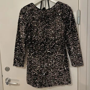 Svart paljettklänning med rosett - Glittrig svart kortklänning täckt med paljetter och en snygg rosett i nacken. Klänningen har långärmade ärmar och en rundad halsringning. Perfekt för dig som vill sticka ut med en festlig look och älskar detaljer som glittrar.