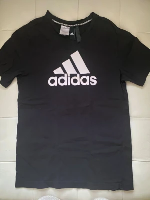 Svart Adidas t-shirt med logga, L - Svart t-shirt från Adidas med stor vit logga framtill. Klassisk rund halsringning och korta ärmar. Tillverkad i mjuk bomull, perfekt för en sportig och avslappnad stil. Snygg detalj med text på insidan av kragen. Det står L i den, men den känns mindre.. (om jag ska vara ärlig) L och M blir bra