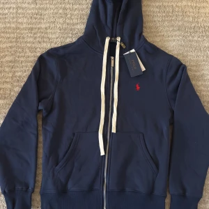Marinblå hoodie från Polo Ralph Lauren - Snygg marinblå hoodie från Polo Ralph Lauren med röd broderad logga på bröstet. Tröjan har huva med vita snören, dragkedja framtill och två stora fickor. Tillverkad i mjuk bomull, perfekt för chill dagar. Märkeslappen sitter kvar.