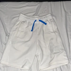 Vita Nike shorts med blå snörning - Nike shorts i vitt med elastisk midja och blå snörning. Snyggt diskret Nike Swoosh-tryck och TM-detalj på benet. Två fickor framtill och mjukt material som känns skönt mot huden. Perfekta för sommaren eller träning.