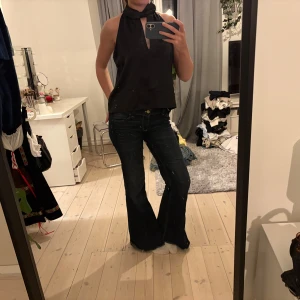 Zara svart topp - Säljer en svart halterneck topp med omlott-detalj vid halsen och en snygg öppning framtill. Toppen är ärmlös och har en loose passform, perfekt för dig som gillar stilrena och trendiga plagg. Materialet känns lent och har ett lätt glansigt utseende.