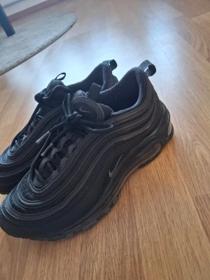Svarta Nike Air Max 97 sneakers 37.5 - Säljer ett par svarta Nike Air Max 97 sneakers med ikoniska vågiga linjer och synlig luftdämpning i sulan. Skorna har snörning, rund tå och är tillverkade i syntet och mesh för en sportig look. Perfekta för dig som gillar streetstyle och vill ha något som sticker ut.