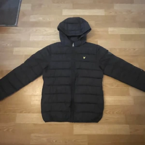Svart pufferjacka från Lyle & Scott - Svart pufferjacka från Lyle & Scott med huva och dragkedja framtill. Jackan har ett quiltat mönster och den klassiska Lyle & Scott-loggan i gult på bröstet. Perfekt för kalla dagar och har en stilren look som funkar till det mesta.