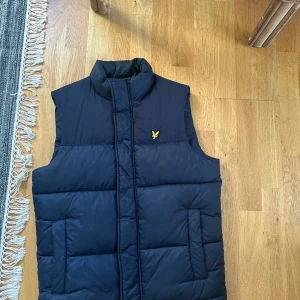 Mörkblå dunväst från Lyle & Scott - Snygg mörkblå dunväst från Lyle & Scott med gul logga på bröstet. Västen har hög krage, två sidofickor och stängs med dragkedja och knappar. Perfekt för lager-på-lager och har en quiltad look som ger extra värme. Materialet är polyester.