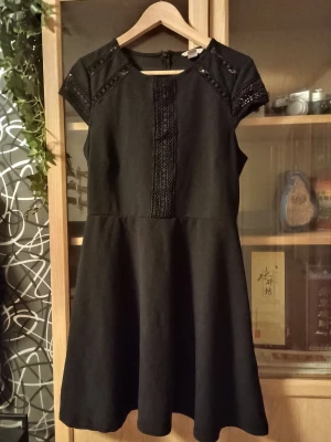 Svart kortklänning med spetsdetaljer - Snygg svart klänning med korta ärmar och spetsdetaljer på axlar och längs bröstet. Modellen är A-linjeformad med rund hals och dragkedja i ryggen. Perfekt för dig som gillar stilrena plagg med en twist. Materialet känns mjukt och har lite struktur. Slutar strax ovanför knäet. Storlek är Medium.