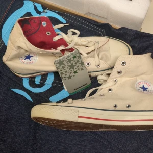 Converse All Star vita high tops - Säljer ett par klassiska Converse All Star high top sneakers i vit textil med röda och blå detaljer. Skorna har snörning, rund tå och platt sula. Insidan är röd med Converse-logga, yttersulan är i gummi. Perfekt för dig som gillar retrostil och streetwear.