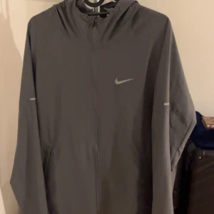 Nike Miller - Säljer en grå vindjacka från Nike med huva och hel dragkedja framtill. Jackan har lång ärm och diskret Nike-logga på bröstet. Perfekt för dig som gillar sportig stil och vill ha något lätt att slänga på sig när det blåser.