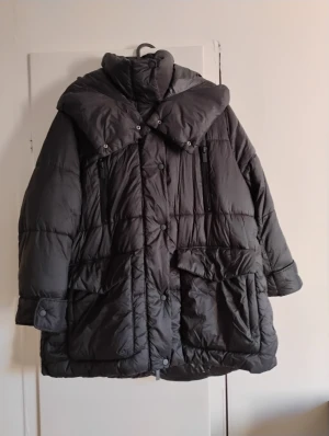 Svart pufferjacka från Zara - Säljer en svart pufferjacka från Zara med stor krage och huva. Jackan har knappar och dragkedja framtill, samt stora fickor med lock. Den är quiltad och har långärmade ärmar med knappdetalj vid mudden. Perfekt för kalla vinterdagar.