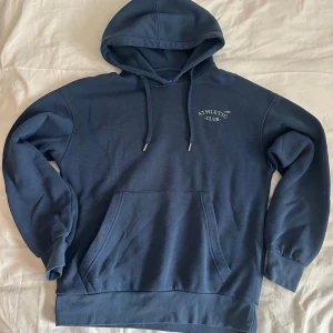 Mörkblå hoodie Athletic Club M - Mörkblå hoodie från Athletic Club med broderad vit text på bröstet. Klassisk känguruficka, huva med snörning och mjuk fleece på insidan. Perfekt för dig som gillar en enkel och stilren look. Pris diskuteras vid snabb affär! 💕💕