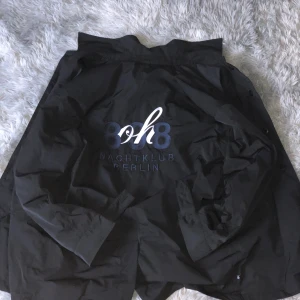 Svart vindjacka Adidas x 808 Berlin - Svart vindjacka från Adidas i samarbete med 808 Nachtklub Berlin. Jackan har broderad logga och text på ryggen, tryckknappar framtill och långärmad design. Materialet är lätt och består av polyester, perfekt för dig som gillar streetwear och klubbkänsla.