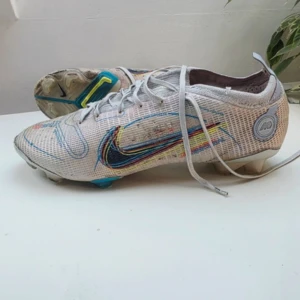 Nike fotbollsskor med färgglada detaljer - Säljer ett par vita Nike fotbollsskor med färgglada detaljer och broderad Swoosh i blått, gult och rött. Skorna har snörning, platt sula med dobbar och är tillverkade i syntetmaterial. Perfekta för dig som vill sticka ut på planen!