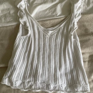 Vit stickad linnetopp från ONLY - Supersöt vit stickad linnetopp från ONLY med volangdetaljer vid axelbanden och v-ringad hals. Toppen har ett ribbat mönster och är ärmlös, perfekt för varma dagar. Materialet är mjukt och luftigt, vilket gör den skön att bära.