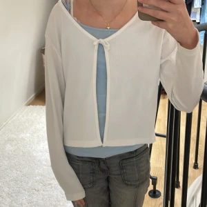 Vit tunn kofta med rosett - Superfin vit topp från H&M med ång ärm. Toppen har en öppen front med söt knytning upptill och croppad passform. Perfekt att slänga över ett linne eller klänning för en trendig lager-på-lager-look.