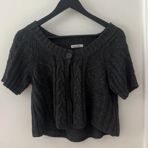 Svart stickad kofta med kort ärm - Svart stickad kofta från Crossroads med korta ärmar och grovt kabelstickat mönster. Koftan har en stor ribbad krage, knapp framtill och en loose passform. Perfekt för lager-på-lager och chill höst/vinter vibes.