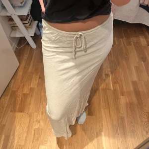 Beige långkjol med snörning - Säljer en beige långkjol med dragsko i midjan. Kjolen har en rak och avslappnad passform och är tillverkad i ett mjukt material som känns skönt mot huden. Perfekt för dig som gillar en enkel och stilren look.