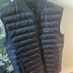 Svart dunväst från Gant - Snygg svart dunväst från Gant med quiltad design och dragkedja framtill. Västen har en stilren look med diskret logga på bröstet och ribbad krage. Perfekt att slänga över en hoodie eller tröja för extra värme under kyliga dagar. Anväds max 2-3 gånger. Original pris 1400