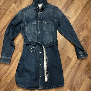 Jeansklänning från Calvin Klein - Snygg och tidlös jeansklänning/skjortklänning från Calvin Klein med långa ärmar, två bröstfickor och knappar framtill. Klänning med klassisk skjortkragen och  ett avtagbart bälte som antingen bärs med blåa sidan ut eller med loggan ut. Perfekt för dig som gillar en cool och avslappnad stil.  Snygg att styla både till vardags och fest – funkar lika bra med sneakers som boots och klackar. Mycket sparsamt använd och för mig inga synliga fel