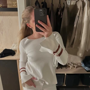 Vit tröja med volangärmar - Mjuk vit tröja med volangärmar och två bruna ränder vid ärmsluten. Rund halsringning och knytband vid båda ärmarna som ger en elegant detalj. Från YAYA woman