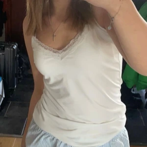 Vit linnetopp med spetskant - Säljer en söt vit linnetopp med smala axelband och fin spetskant längs v-ringningen. Toppen är i ett mjukt och glansigt material som känns lyxigt mot huden. Perfekt att styla med jeans eller kjol för en fräsch look.