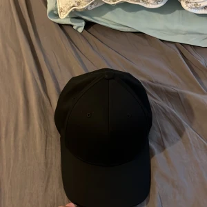 Svart keps från Flexfit Youth.  - Säljer en stilren svart keps från Flexfit i ungdomsstorlek. Kepsen har böjd skärm med vit undersida och klassisk sexpanelsdesign. Tillverkad i ett mjukt och flexibelt material som sitter skönt på huvudet. Perfekt för en clean och enkel look.