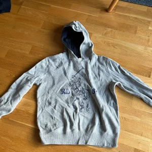Grå hoodie från Polo Ralph Lauren - En riktigt snygg hoodie från polo Ralph Lauren. Storleken är ungefär för 12-13 åringar kan även funka för 11 åringar beroende på längd den har krympt en del men funkar ändå Perefekt. Den har ett litet hål under den högra armhålan (se bild 3) men syns knappt. Vid minsta lilla fråga kan du skriva till mig.