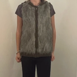 Grå pälsväst med huva och dragkedja - Snygg grå pälsväst med stickad baksida och huva. Framsidan är täckt av mjuk fuskpäls och västen stängs med dragkedja. Perfekt för lager-på-lager och ger en cool look till din outfit.