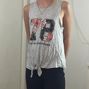 Grå ärmlös linnetopp med tryck - Säljer en grå ärmlös linnetopp med stort tryck '78' i blommigt mönster och texten 'YOU ARE UNSTOPPABLE' under. Toppen har en snygg knytning framtill som ger en chill vibe. Perfekt för dig som gillar sportig och avslappnad stil.