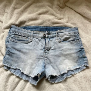 Ljusblå jeansshorts från H&M - Snygga ljusblå jeansshorts från H&M med råa, fransiga kanter och klassisk femficksdesign. Shortsen har dragkedja och knapp framtill samt bälteshällor. Perfekta för varma dagar och en avslappnad look.