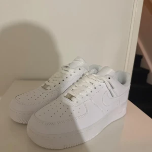 Nike Air Force 1 helvita sneakers - Säljer ett par helt nya Nike Air Force 1 i helvitt skinn. De har låg profil, rund tå, snörning och den ikoniska swoosh-loggan på sidan. Perfekta för dig som vill ha en clean och tidlös look. Passar till allt och är superenkla att matcha.