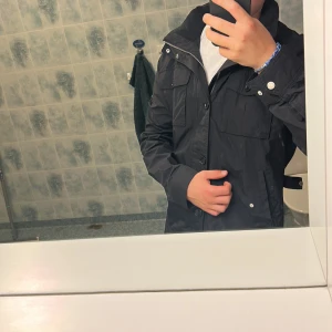 Svart vindjacka från J.Lindeberg - Snygg svart vindjacka från J.Lindeberg med flera fickor framtill, dragkedja och knappar. Jackan har en klassisk krage och cool metallbadge på ärmen. Perfekt för dig som vill ha en stilren och funktionell jacka med streetkänsla. som ny