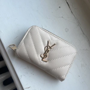 Beige quiltad plånbok från YSL - Snygg beige plånbok från YSL med quiltad design och guldigt YSL-emblem på framsidan. Plånboken har dragkedja runtom och flera fack inuti för kort och mynt. Materialet är mjukt skinn och detaljerna är i guldmetall. Perfekt storlek för att ha med sig överallt. vet inte riktigt om den är äkta eftersom jag fick den av min farmor