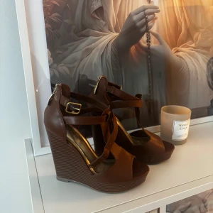 Bruna kilklackar med remmar - Snygga bruna kilklackar från Guess med korsade remmar och öppen tå. Skorna har justerbart spänne i guld och hög plattformsklack. Perfekta för dig som vill ha både höjd och komfort i en stilren design. Dessa har aldrig varit använda! Pris går att pruta.