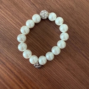 Pärlarmband med Gemini-hänge - Stilrent armband med stora vita pärlor och en glittrig silverfärgad kula. Armbandet har ett hjärtformat stjärntecken-hänge i silver med texten Gemini. (Töjbart och stretchigt så passar flera storlekar). Okänt vart armbandet är köpt.