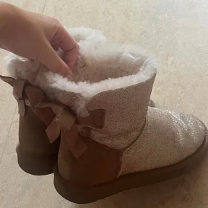 Glittriga beige boots med rosett - Supermysiga boots i beige med glittrig ovandel och fluffigt vitt foder. Skorna har rund tå, platt sula och fina rosetter bak. Perfekta för kalla dagar när du vill vara både varm och snygg.