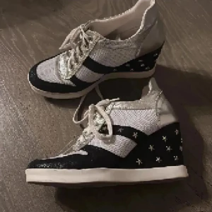 Silvriga sneakers med kilklack och stjärnor - Unika sneakers från Heine i silver och svart med glittriga detaljer och kilklack. Skorna har snörning, svarta ränder och små stjärnor på klacken. Materialet är syntet med metallic-finish och vit sula. Perfekta för dig som vill sticka ut med din stil.
