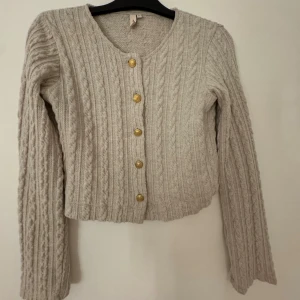 Beige stickad kofta från Nelly - Supermysig beige stickad kofta från Nelly i storlek XS. Koftan har ett snyggt kabelstickat mönster, rund hals och guldiga knappar framtill. Långärmad och perfekt att slänga på sig när det är lite kyligt ute.