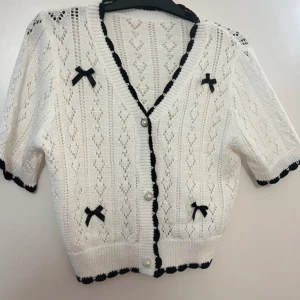 Vit stickad kofta med svarta rosetter - Supersöt vit stickad kofta med korta ärmar, svarta rosetter och pärlknappar. Koftan har svarta detaljer längs kanterna och ett fint hålmönster över hela plagget. Perfekt för dig som gillar gulliga och unika detaljer.