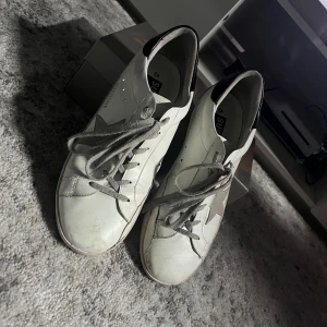 Golden Goose Superstar  - Golden Goose Superstar sneakers i vitt skinn med grå mockadetaljer och grå snörning. Klassisk stjärna på sidan och svart hälparti. Snygg och tidlös modell som passar till det mesta. Levereras med originalkartong.