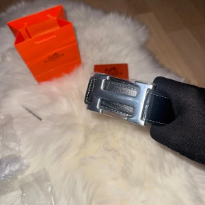 Svart  skärp med H-spänne - Snyggt svart bälte från H med klassiskt H-spänne i silverfärgad metall. Bältet är gjort av skinn med vit kontrastsöm längs kanterna och har en stilren, rak form. Perfekt för att lyfta din outfit med en lyxig touch.