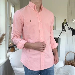 Ljusrosa skjorta från Ralph Lauren - Säljer en snygg ljusrosa skjorta från Ralph Lauren med klassisk button-down krage och broderad logga på bröstet. Skjortan är långärmad, har normal passform och är tillverkad i mjuk bomull. Perfekt för dig som gillar stilrena och fräscha plagg.