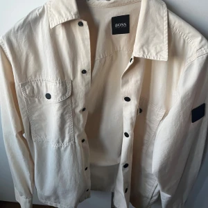 Beige overshirt från BOSS - Säljer en stilren beige overshirt från BOSS med svarta knappar och två bröstfickor med lock. Jackan har klassisk krage och patch på ärmen. Perfekt att slänga över en hoodie för en clean look. Kontakta gärna vid intresse😊