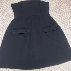 Svart axelbandslös kort klänning - Snygg svart axelbandslös klänning med kort längd och två dekorativa ficklock framtill. Stilren och enkel design som ger en chic look. Perfekt för dig som gillar minimalistiska plagg med en twist.